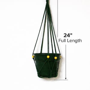 Könnte beinhalten: Ein dunkelgrüner Hängepflanzentopf mit einer Gesamtlänge von 61 cm. Der Topf wird von dunkelgrünen Kordeln gehalten und hat gelbe Perlen. Der Topf ist für eine kleine Pflanze konzipiert und an einem Metallring befestigt.