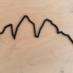 Può includere: Contorno nero di una catena montuosa su uno sfondo chiaro con venature del legno. Il design geometrico semplice presenta picchi e valli. Probabilmente si tratta di un oggetto decorativo o di un'opera d'arte murale.