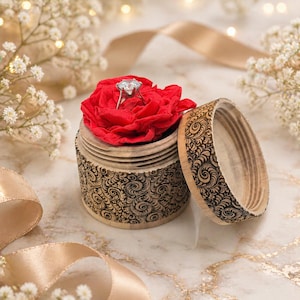 Può includere: Un anello di fidanzamento con diamanti è posto in una rosa rossa all'interno di un portagioie in legno aperto e finemente intagliato. La scatola e il coperchio presentano un motivo a spirale dettagliato. Illuminazione soffusa e accenti floreali.