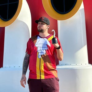Op de afbeelding: Een persoon draagt een rood, geel en wit shirt met een piratenontwerp en een zwarte pet staat op een houten dek voor een rood, wit en blauw schip. Het schip heeft een groot Mickey Mouse-hoofd aan de voorkant.