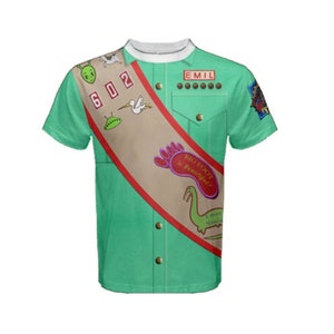 Może przedstawiać: Zielony T-shirt z krótkim rękawem, brązową szarfą i naszywkami. Na szarfie widnieje napis "BIG FOOT is Beautiful!" a naszywki przedstawiają obrazy UFO, ptaka, śladu stopy i dinozaura. T-shirt ma biały kołnierzyk i guziki z przodu. Na koszulce widnieje napis "EMIL" i "602".