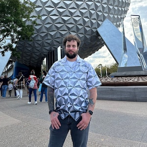 May include: A person wearing a white and gray patterned shirt stands in front of a large, silver geodesic dome. The dome is a popular landmark and tourist attraction.