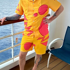 May include: A man wearing a yellow and red patterned shirt and matching shorts with a cartoon character design. He is wearing a black hat with ears and blue sunglasses. He is standing on a cruise ship balcony.