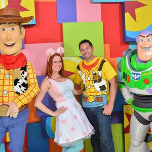 May include: A costumed Woody character, a person in a pink and white polka dot dress with mouse ears, a person in a yellow Woody-themed t-shirt and blue jeans, and a costumed Buzz Lightyear character standing in front of a colorful block wall.