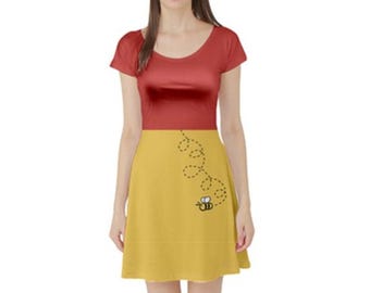 Pode incluir: Um vestido de manga curta com um corpete vermelho liso de decote redondo e uma saia amarelo-mostarda. A saia apresenta um pequeno gráfico de uma abelha com uma linha preta tracejada que descreve uma trajetória de voo em espiral ascendente.