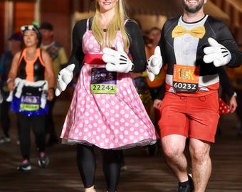 May include: A couple dressed in Mickey Mouse and Minnie Mouse costumes are running a race. The woman is wearing a pink polka dot dress with black leggings and pink running shoes. The man is wearing a black and red Mickey Mouse costume with red shorts and black running shoes. Both are wearing race bibs with numbers. The woman's bib reads "22241" and the man's bib reads "60232".