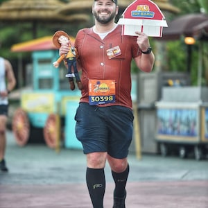 以下が含まれることがあります： 赤と茶色のシャツを着て、漫画のキャラクターがプリントされた男性がレースをしています。彼は「Al's Toy Barn」と書かれた看板と、トイストーリーのウッディのぬいぐるみを持っています。彼は30398のレースナンバーをつけています。