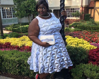 Puede incluir: Una mujer con un vestido blanco y gris con estampado geométrico y un bolso blanco que dice "Spaceship Earth". Está de pie frente a un jardín de flores rojas, amarillas y verdes.