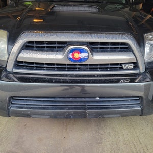 May include: Black SUV with a silver grill and a Colorado state flag emblem. The grill has the text "V8" on it. The vehicle's headlights are visible, and the front bumper is dark gray.