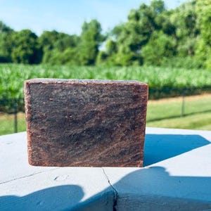 May include: A rectangular bar of soap with a dark brown color and a textured surface. The soap is sitting on a white surface with a green field and trees in the background.
