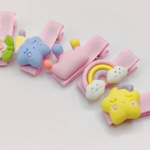 May include: Set of six pink hair clips with colorful cartoon characters. The characters include a flower, a star, a crown, a rainbow, a cloud, and a star.