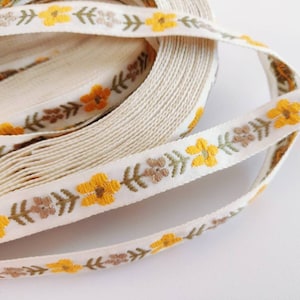 May include: A roll of white cotton ribbon with a yellow floral pattern. The ribbon has a repeating pattern of small yellow flowers and green leaves.