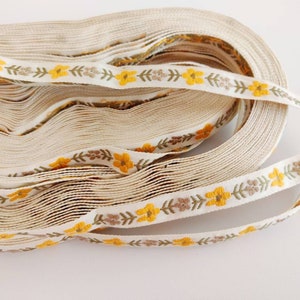 May include: A roll of white cotton ribbon with a repeating pattern of yellow flowers, brown leaves, and green stems.