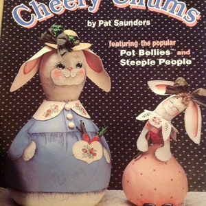 Puede incluir: Una portada de libro con dos calabazas pintadas con forma de conejos. Las calabazas están vestidas con vestidos azules y rosas con detalles blancos. El título del libro es "Cheery Chums" y el autor es Pat Saunders. El libro presenta los populares Pot Bellies y Steeple People.