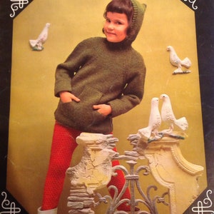 May include: A young girl wearing a green knitted sweater with a hood and red pants stands in front of a white stone wall with a wrought iron gate. The girl is looking at the camera. There are white ceramic doves on the wall.