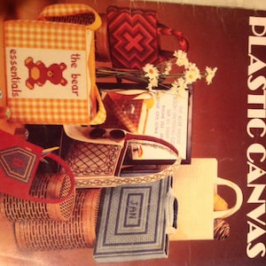 May include: A book cover with the title "Needlepoint Totes for Plastic Canvas" and a picture of various needlepoint totes, including a red tote with a diamond pattern, a yellow tote with a bear design, and a blue tote with the word "Jan" on it.