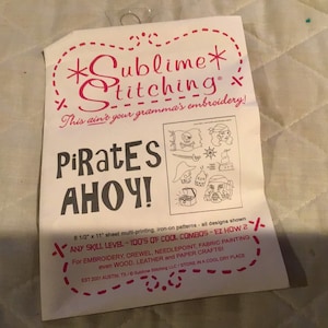 Op de afbeelding: Een wit vel papier met zwarte en roze tekst die "*Sublime* Stitching This ain't your gramma's embroidery! Pirates AHOY!" luidt. Het papier bevat ook een zwart-witte illustratie van een piratenschip, een schatkist en een piratengezicht. De tekst onderaan luidt "8 1/2" x 11" sheet multi-printing, iron-on patterns - all designs shown ANY SKILL LEVEL - 100'S OF COOL COMBOS - EZ HOW 2 For EMBROIDERY, CREWEL, NEEDLEPOINT, FABRIC PAINTING even WOOD, LEATHER and PAPER CRAFTS! EST 2001 AUSTIN, TX / Sublime Stitching LLC / STORE IN A COOL DRY PLACE"
