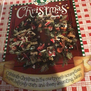 May include: A Gooseberry Patch Christmas Book 6 softcover book with a red and white plaid background. The cover features a green wreath decorated with ornaments and small wooden blocks with text, and a banner with the text Celebrate Christmas in the country with scrumptious recipes, holly-jolly crafts and cheery decorating ideas!