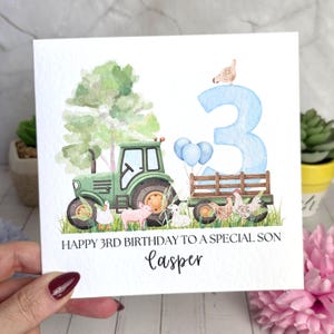May include: A square greeting card featuring a watercolour illustration of a green tractor pulling a trailer, a large light blue number 3, farm animals including a pig, sheep, and chickens, and the text HAPPY 3RD BIRTHDAY TO A SPECIAL SON Casper.