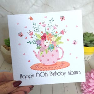 May include: A square greeting card with a white background featuring a pink polka-dot teapot filled with colorful flowers and green leaves. Small butterflies and hearts surround the teapot, which sits on an orange mat. Text reads: Happy 60th Birthday Mama.