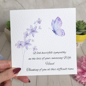 Può includere: Un biglietto di condoglianze quadrato con un'illustrazione ad acquerello di fiori viola chiaro e una farfalla abbinata. Il testo recita: With heartfelt sympathy on the loss of your amazing Wife Hazel. Thinking of you at this difficult time.