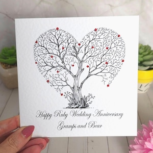 Può includere: Un biglietto d'auguri quadrato bianco con un'illustrazione a inchiostro nero di un albero a forma di cuore. I rami dell'albero sono decorati con piccole gemme rosse. Il testo recita: Happy Ruby Wedding Anniversary, Gramps and Bear, in corsivo nero.
