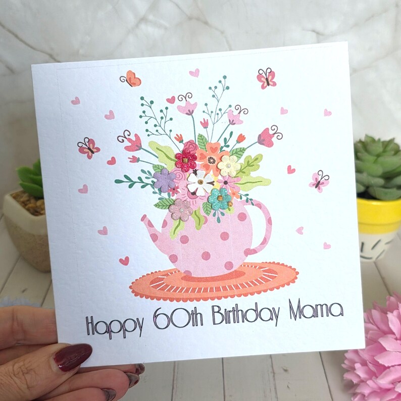 May include: A square greeting card with a white background featuring a pink polka-dot teapot filled with colorful flowers and green leaves. Small butterflies and hearts surround the teapot, which sits on an orange mat. Text reads: Happy 60th Birthday Mama.