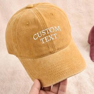 Puede incluir: Gorra de béisbol de color amarillo mostaza con el texto "CUSTOM TEXT" bordado en blanco. La gorra tiene una visera curva y un diseño clásico de seis paneles. Una segunda gorra granate es visible en el fondo.