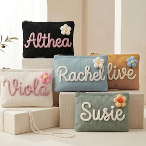 May include: Five rectangular corduroy zipper pouches in black, cream, light blue, mustard yellow, and sage green. Each pouch features a raised, cursive name in white or pink yarn and a small, colorful crocheted flower accent attached to the front.