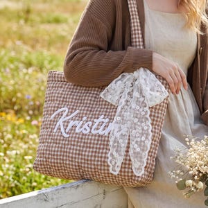 May include: A quilted brown and white gingham tote bag featuring the name Kristin embroidered in white script lettering on the front, accented with a decorative white lace ribbon tied to the shoulder strap.