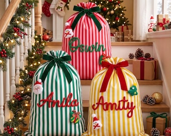 Op de afbeelding: Drie stoffen kerstcadeauzakken met verticale strepen in rood, groen en geel. Elke zak is voorzien van een fluwelen strik aan de bovenkant, een pluche kerstornament en een in cursief geborduurde naam: Oswin, Anila en Amity.