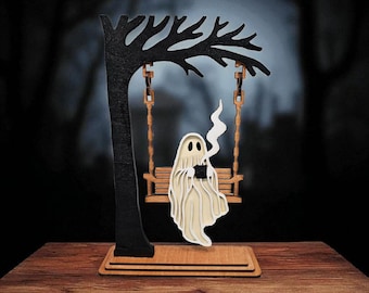 May include: A wooden Halloween decoration featuring a ghost on a swing. The ghost, holding a cup, is made of layered beige wood, with a wisp of smoke rising. The swing hangs from a black tree, all set on a wooden base.