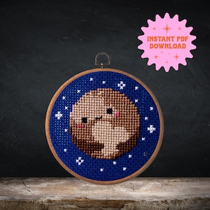 Könnte beinhalten: Ein Kreuzstich-Stickrahmen mit blauem Hintergrund und weißen Sternen. In der Mitte des Rahmens befindet sich ein braunes und beigefarbenes Kreuzstich-Design eines Planeten mit einem lächelnden Gesicht.