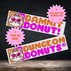 Può includere: Due schemi a punto croce con un gatto pixelato che indossa una corona e occhiali da sole. Il motivo superiore riporta la scritta DAMMIT DONUT! in arancione e rosa, quello inferiore riporta DUNGEON DONUTS in arancione e rosa. Include etichette rosa.