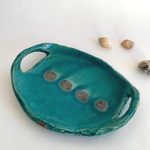 Op de afbeelding: Een handgemaakte, turquoise keramische schaal met twee handvatten. De ovale schaal heeft vier donkerbruine, cirkelvormige accenten. De schaal staat op een wit oppervlak, met verschillende schelpen op de achtergrond.