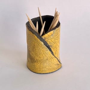 Może przedstawiać: Ceramiczny pojemnik na wykałaczki w kolorze żółtym z czarnym szkliwem craquelé. Pojemnik jest cylindryczny i ma lekko nierównomierny kształt. Jest wypełniony drewnianymi wykałaczkami.