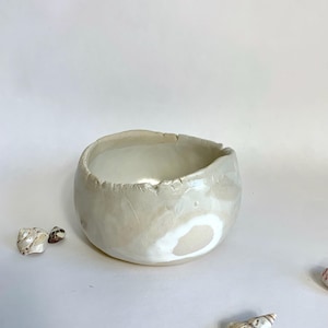 Könnte beinhalten: Eine kleine, cremefarbene Keramikschale mit strukturierter Oberfläche und leicht unregelmäßigem Rand. Die Schale hat eine runde Form und ein glattes Inneres. Mehrere Muscheln sind um die Schale herum verstreut, was auf ein Küstenmotiv hindeutet.