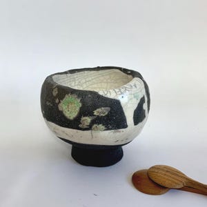 Könnte beinhalten: Eine handgefertigte Keramikschale mit einem abstrakten schwarzen, weißen und grünen Design. Die Schale hat eine runde Form mit einem kleinen Sockel. Zwei kleine Holzlöffel liegen neben der Schale.