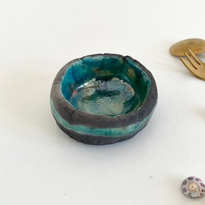 Può includere: Un piccolo piatto rotondo in ceramica con una glassa turchese e strisce nere. Il piatto è fatto a mano e ha un aspetto rustico e organico.