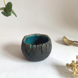 Peut inclure: Un petit bol rond en céramique avec un extérieur gris foncé texturé et un intérieur bleu sarcelle vif. Le bol a une apparence craquelée et vieillie. Quelques brins de verdure et une fourchette dorée sont en arrière-plan.