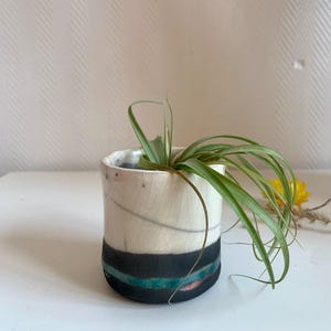 Op de afbeelding: Een kleine, handgemaakte keramische plantenbak met een witte, gebarsten glazuur. De plantenbak heeft een zwarte band met turquoise en koperkleurige strepen. Er zit een groene luchtplant in. De plantenbak staat op een wit oppervlak.