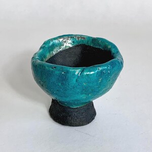 Peut inclure: Un bol en céramique fait à la main, extérieur turquoise et intérieur noir. Le bol a une surface texturée et repose sur une base sombre et texturée. Le bord du bol a une forme organique et irrégulière.