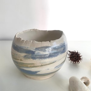 Può includere: Una ciotola in ceramica fatta a mano con un bordo ruvido. La ciotola presenta pennellate orizzontali blu, verdi e crema. Un oggetto decorativo per la casa.