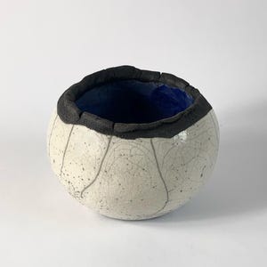 May include: A small white ceramic bowl with a black rim and a blue glaze inside. The bowl has a crackled glaze and a textured surface.