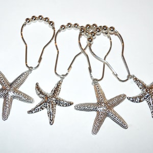 May include: Set of four silver-colored shower curtain hooks. Each hook features a starfish charm. The starfish are detailed with textured surfaces and attached to the hooks with a small loop. The hooks have a beaded design.