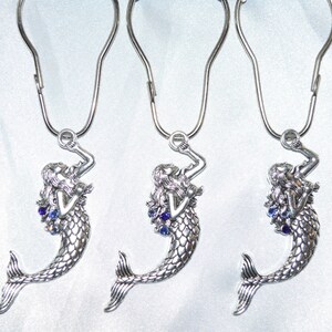 Puede incluir: Tres ganchos plateados para cortina de ducha con forma de sirena y detalles de gemas azules. Cada figura de sirena tiene cabello suelto y una cola escamada, suspendida de un gancho de metal curvado.