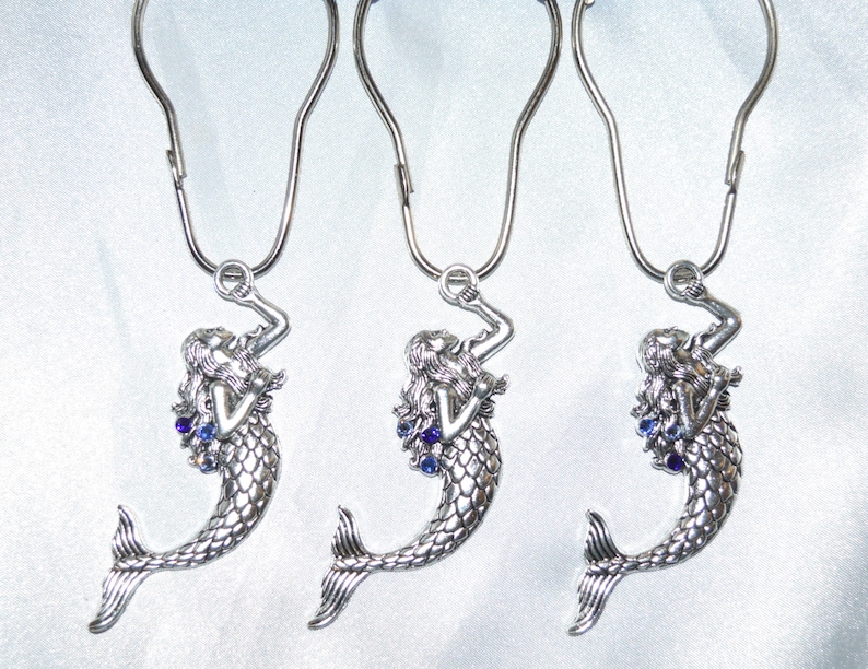 Puede incluir: Tres ganchos plateados para cortina de ducha con forma de sirena y detalles de gemas azules. Cada figura de sirena tiene cabello suelto y una cola escamada, suspendida de un gancho de metal curvado.