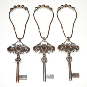 May include: Three silver-colored shower curtain hooks. Each hook features a decorative key pendant with ornate detailing. The hooks have a loop of small silver beads at the top, designed to hang from a shower rod. The keys have a vintage, antique aesthetic.