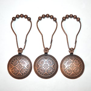 May include: Three bronze shower curtain hooks. Each hook features a circular, decorative medallion with an embossed floral pattern. The hooks have a looped top with small ball accents, and are designed to hang a shower curtain.