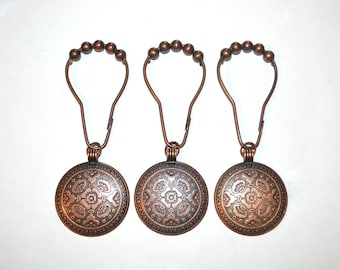 Puede incluir: Tres ganchos de cortina de ducha de bronce. Cada gancho presenta un medallón circular decorativo con un patrón floral en relieve. Los ganchos tienen una parte superior en forma de bucle con pequeños detalles de bolas, diseñados para colgar una cortina de ducha.
